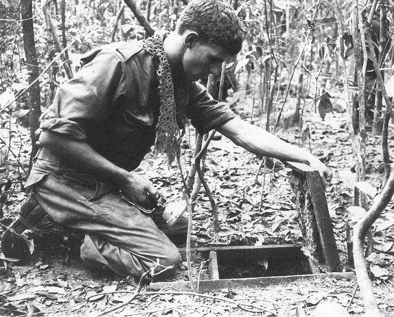 A soldier is kneeling over an open trapdoor on the jungle floor while holding a torch. An Australian sapper inspects a Viet Cong tunnel discovered during Operation Crimp, South Vietnam 1966